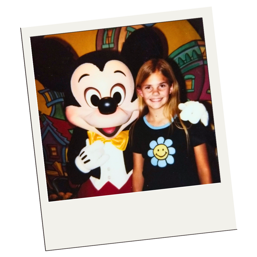 A Polaroid style photo of me with Mickey Mouse. Mickey has his arm around me and I am wearing a black t-shirt with a yellow and blue daisy smiley face flower.